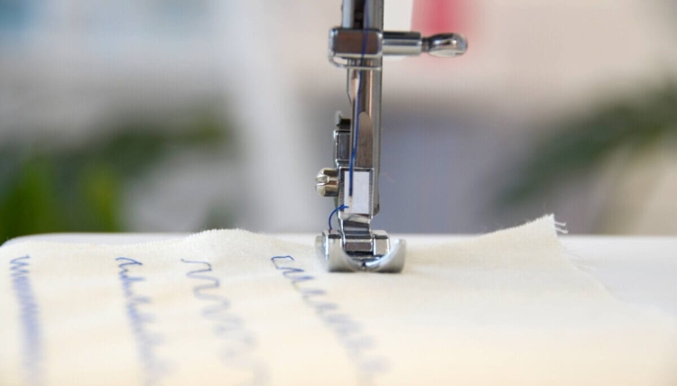 Maszyna do szycia BERNETTE Sew&Go 1 w zbliżeniu, z widoczną stopką i igłą szyjącą materiał. Niebieska nici na jasnej tkaninie, pokazująca ścieg dekoracyjny. Tło rozmyte, co podkreśla detale maszyny i materiału.