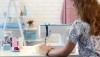 Maszyna do szycia BERNETTE Sew&Go 1 w kolorze białym z niebieskimi akcentami, mechaniczna z widocznymi 10 ściegami. Ujęcie z boku, produkt stoi na stole, użytkownik trzyma materiał do szycia. W tle akcesoria krawieckie oraz pastelowe elementy dekoracyjne