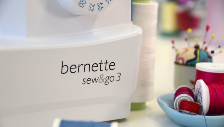 Maszyna do szycia BERNETTE Sew&Go 3 w kolorze białym z widocznym logo. Ujęcie z boku, pokazujące pokrętło z różnymi ściegami. W tle szpule nici w kolorach czerwonym, białym i zielonym oraz pin pinowy.