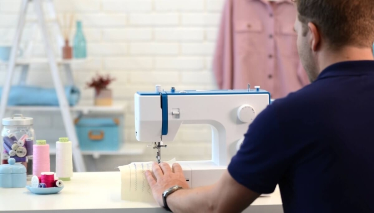 Maszyna do szycia BERNETTE Sew&Go 3 w kolorze białym z niebieskimi akcentami, mechaniczna, z widocznym ząbkowanym ściegiem na tkaninie. Ujęcie z boku, mężczyzna trzyma materiał, w tle szafa z kolorowymi akcesoriami krawieckimi.