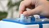Maszyna do szycia BERNETTE Sew&Go 5 z białą obudową i niebieskimi detalami. Widoczna ręka obracająca pokrętło do ustawiania ściegów, z oznaczeniami od 1 do 4. Na górze maszyny umieszczono także inne elementy sterujące oraz igłę. Tło rozmyte.