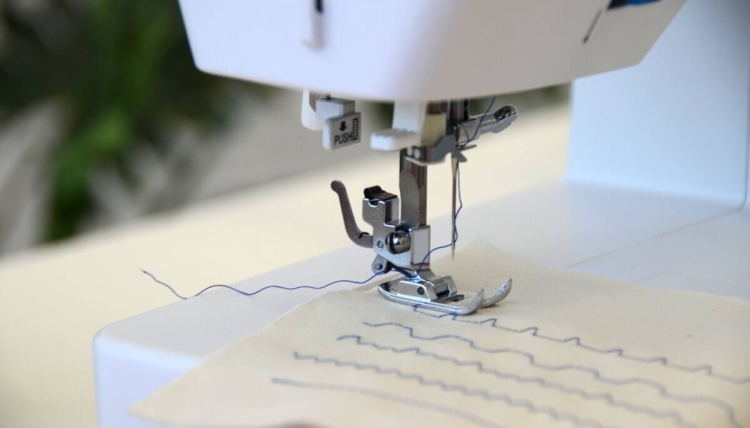 Maszyna do szycia BERNETTE Sew&Go 5 w zbliżeniu, z widoczną stopką, igłą oraz niebieską nicią szyjącą. Na materiale widać równo ułożone ściegi, a całość umieszczona na jasnym tle.