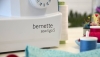Maszyna do szycia BERNETTE Sew&Go 5 w kolorze białym, z pokrętłem do wyboru 23 ściegów. Widoczne logo bernatette sew&go 5. W tle kolorowe szpule nici oraz igły w pin holderze. Produkt umieszczony na jasnym stole, w zbliżeniu na detale.