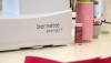 Maszyna do szycia BERNETTE Sew&Go 7 w kolorze białym z widocznym logo. Obok znajdują się szpule nici w odcieniach różu i fioletu oraz pojemnik z igłami. Ujęcie z boku, ukazujące detal maszyny i akcesoria do szycia.