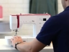 Maszyna do szycia BERNETTE Sew&Go 7 w kolorze białym z czerwonymi detalami, z widocznymi przyciskami i wyświetlaczem. Użytkownik trzyma rękę na obszarze szycia, co podkreśla jej ergonomiczny kształt. Produkt stoi na płaskiej powierzchni.