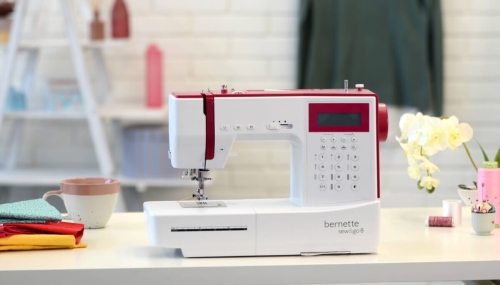 Maszyna do szycia BERNETTE Sew&Go 8 w białym i czerwonym kolorze, frontalnie, z wyświetlaczem i panelem sterującym. W tle widoczne kolorowe materiały, filiżanka oraz kwiaty, co tworzy przyjemną aranżację.