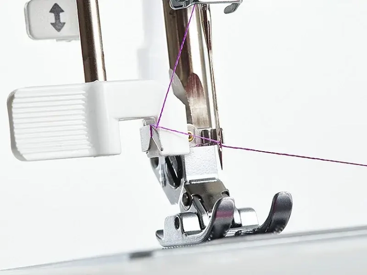 Zbliżenie na igłę maszyny do szycia BERNETTE Sew&Go 8. Widoczne są elementy mechanizmu z białym uchwytem oraz różowa nitka przechodząca przez oczko igły, co ilustruje proces szycia. Tło proste, białe.