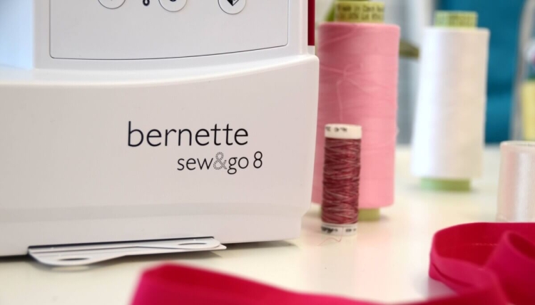 Maszyna do szycia BERNETTE Sew&Go 8 w kolorze białym, z wyraźnym logo. Widoczne 197 ściegów, guziki sterujące. W tle kolorowe szpulki nici: różowe, białe, czerwone, oraz złożony różowy materiał. Ujęcie z boku na stół.
