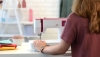 Maszyna do szycia BERNETTE Sew&Go 8 w kolorze białym z różowymi akcentami, z widocznym panelem sterującym, ustawiona na stole. Użytkownik trzyma materiał przy igle. W tle kolorowe kawałki tkanin oraz filiżanka.