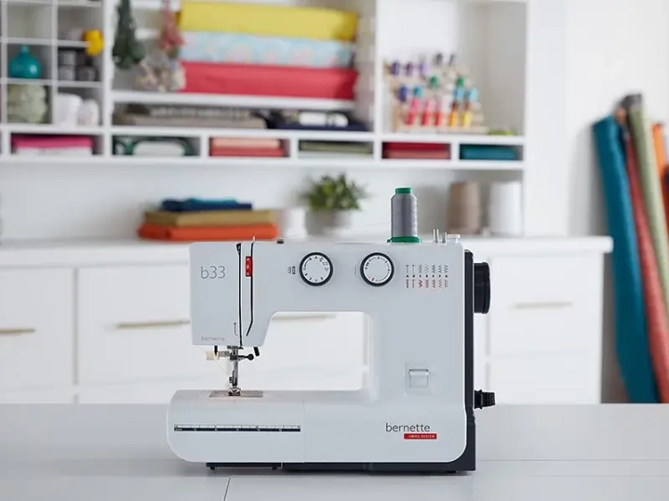Maszyna do szycia BERNETTE B33 w kolorze białym, mechaniczna, z 15 ściegami. Widoczne dwa pokrętła do regulacji oraz szpulka z nicią. Produkt ustawiony na stole, w tle różnokolorowe materiały oraz akcesoria krawieckie.