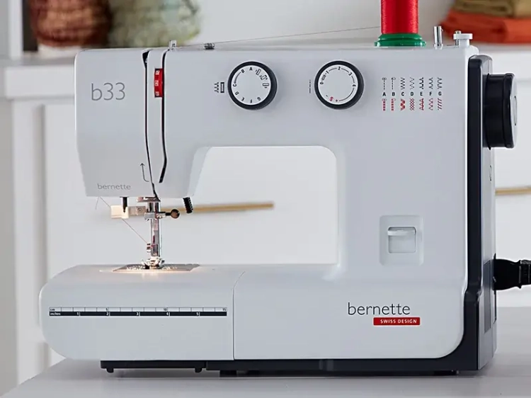 Maszyna do szycia BERNETTE B33 w kolorze białym, z mechanicznymi pokrętłami do ustawiania ściegów i różnymi opcjami ściegów. Widoczne logo bernette oraz oznaczenia funkcji. Ujęcie frontalne, maszyna stoi na płaskiej powierzchni.