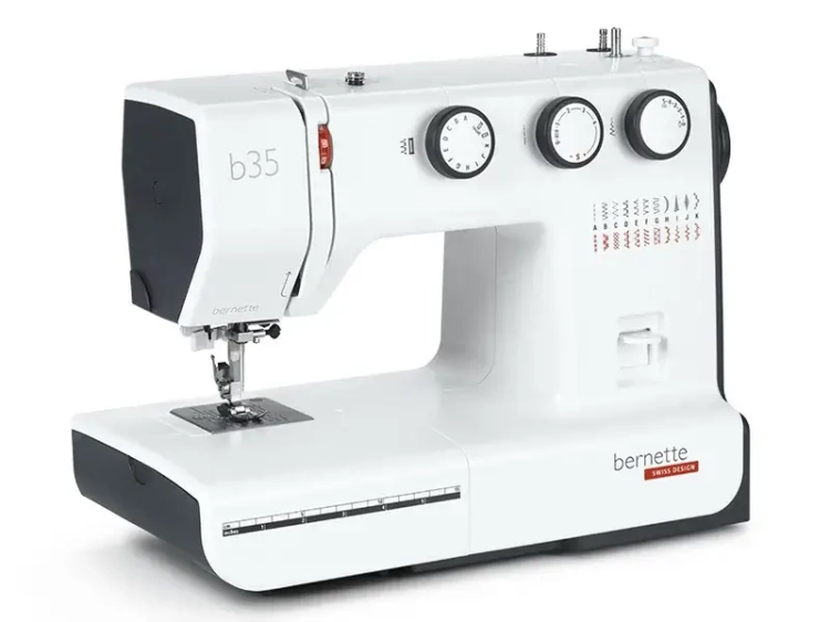Maszyna do szycia BERNETTE B35 w kolorze białym, z mechanicznymi pokrętłami do regulacji oraz 23 ściegami. Ujęcie frontalne, produkt leżący na płaskiej powierzchni, widoczny stół z oznaczeniem długości ściegu i logo marki.