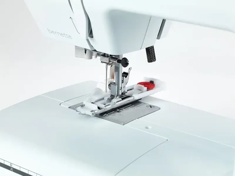 Maszyna do szycia BERNETTE B35 w białej obudowie, z widoczną stopką i igłą. Zastosowanie mechaniczne, oferująca 23 rodzaje ściegów. Zdjęcie zbliżeniowe ukazuje mechanizm szycia oraz miejsce na szpulkę.