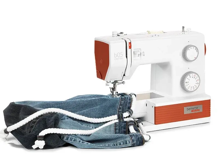 Maszyna do szycia BERNETTE B05 CRAFTER w kolorze białym i pomarańczowym, z 30 ściegami, ustawiona na stole, z kawałkiem materiału dżinsowego z białym sznurkiem. Widziane pokrętła do regulacji ściegów oraz logo marki.