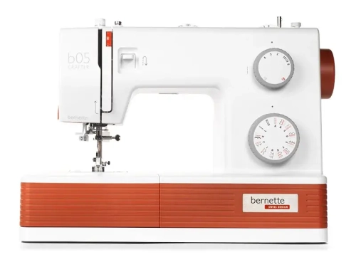 Maszyna do szycia BERNETTE B05 CRAFTER w kolorze białym z pomarańczowym akcentem, z widocznymi pokrętłami do ustawiania ściegów i mechanizmem szyjącym. Ujęcie frontalne, z wyraźnym logo bernette oraz oznaczeniem b05 CRAFTER.