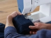 Maszyna do szycia BERNETTE B05 ACADEMY w kolorze niebieskim, z widoczną stopką i materiałem dżinsowym, podczas szycia. Użytkownik trzyma materiał, pokazując mechaniczne ściegi oraz prosty interfejs na maszynie. Tło drewniane.