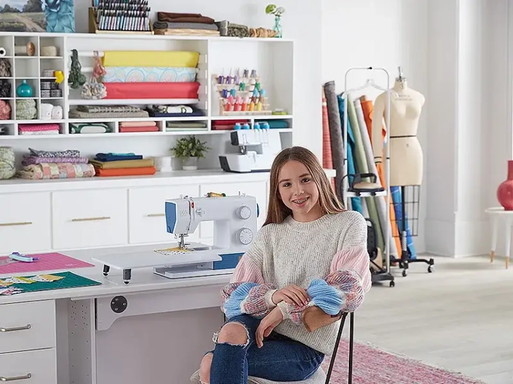 Maszyna do szycia BERNETTE B05 ACADEMY w kolorze białym z niebieskimi detalami, umieszczona na stole. W tle widoczne materiały do szycia, kolorowe nici i manekin. Osoba siedząca obok maszyny, uśmiechnięta, w szarej bluzie.