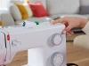 Maszyna do szycia BERNETTE B05 ACADEMY w kolorze białym, z mechanicznymi pokrętłami do wyboru ściegów i ustawień, w zbliżeniu na rękę regulującą ustawienia. W tle widoczne kolorowe poduszki.