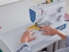 Maszyna do szycia BERNETTE B05 ACADEMY w kolorze białym i niebieskim, z widocznymi pokrętłami do regulacji, szyje materiał w żółtym kolorze z numerami. Ujęcie z bliska, z rękami operatora na tkaninie, na gładkim blacie.
