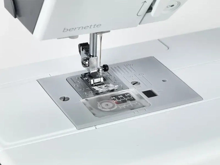 Maszyna do szycia BERNETTE B37 w zbliżeniu, ukazująca stopkę, igłę oraz panel z oznaczeniami ściegów na metalowej podstawie. Widoczne są elementy mechanizmu szycia oraz logo bernette. Tło białe, co podkreśla detale produktu.