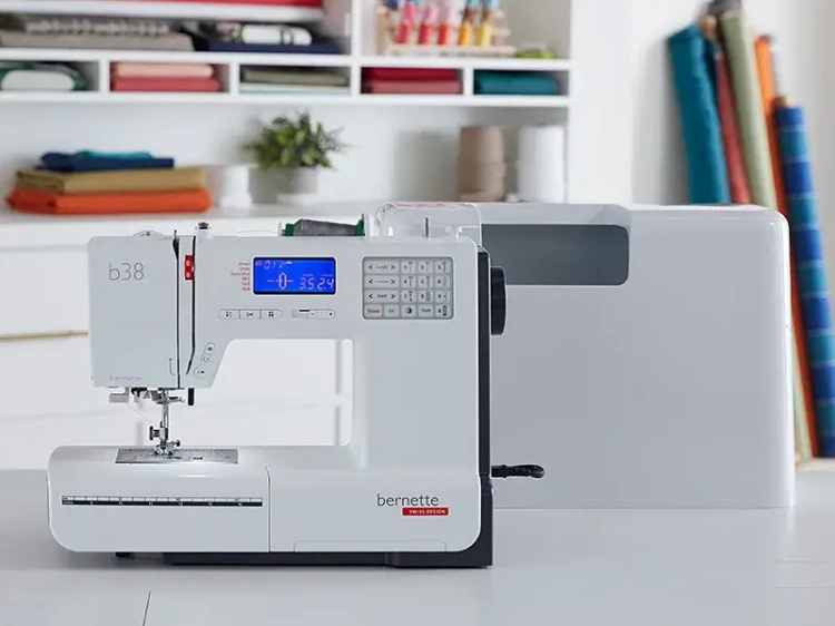 Maszyna do szycia BERNETTE B38 w kolorze białym, z komputerowym panelem sterującym i wyświetlaczem LCD, oferująca 394 różne ściegi. Ujęcie frontalne, z widoczną obudową. Na tle jasnej przestrzeni roboczej z różnokolorowymi materiałami.
