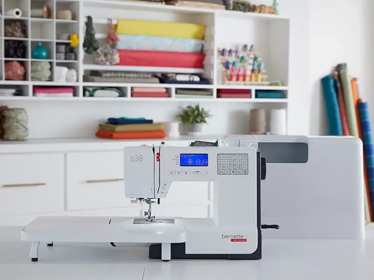Maszyna do szycia BERNETTE B38 w kolorze białym, z dużym wyświetlaczem LCD i 394 ściegami. Ujęcie frontalne, stojąca na stole, z dodatkowym blatem. W tle różnokolorowe materiały i akcesoria krawieckie.