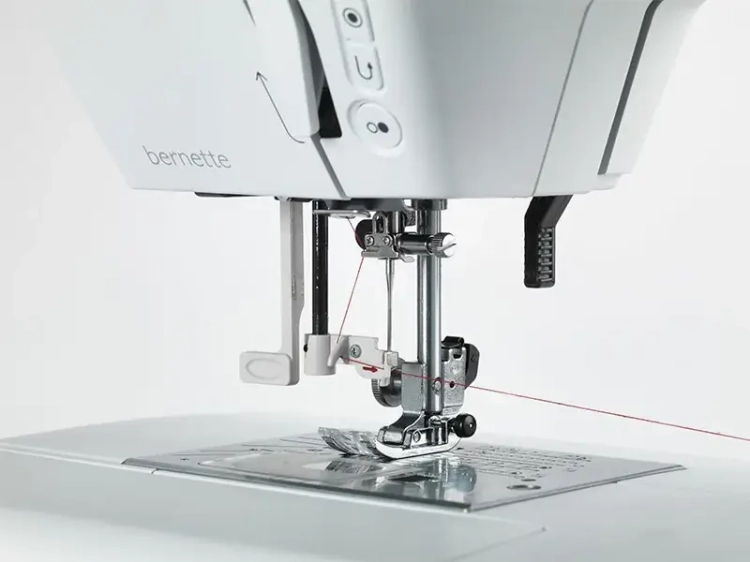 Maszyna do szycia BERNETTE B38 w kolorze białym, z widoczną stopką i igłą. Widać również mechanizm podawania materiału oraz czerwony nit. Ujęcie z bliska, pokazujące detale maszyny i panel sterujący z przyciskami.
