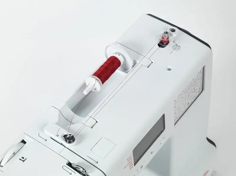 Maszyna do szycia BERNETTE B38, widok z góry. Widać białą obudowę, czerwony szpulę z nicią oraz elementy do nawlekania. Na panelu znajdują się przyciski i wyświetlacz z informacjami o ściegach.