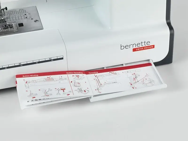 Maszyna do szycia BERNETTE B38 z widoczną szufladą, w której znajduje się instrukcja dotycząca nawlekania i napełniania szpulki. Urządzenie ma białą obudowę z czerwonym logo bernette oraz panel sterujący z różnymi funkcjami.
