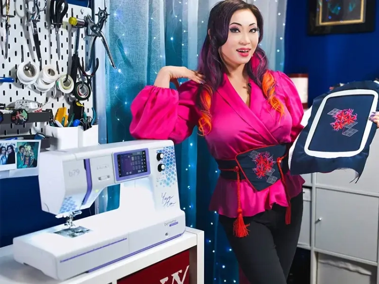 Maszyno-hafciarka BERNETTE B79 SE Yaya Han w kolorze białym z fioletowymi akcentami, z panelem sterującym. Obok osoba trzyma haftowany materiał w ramce, w tle przybory krawieckie na ścianie.