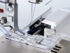 Owerlok BERNETTE B64 AIRLOCK z widocznym stopką i igłą, z nitkami w kolorze niebieskim. Ujęcie z bliska, pokazujące mechanizm szycia oraz przezroczystą stopkę. Elementy metalowe i plastikowe, na szarym tle.