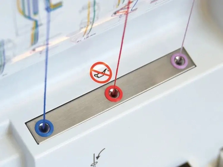 Zbliżenie na stację nawlekania maszyny overlock BERNETTE B68 AIRLOCK. Widoczne trzy nici w kolorach niebieskim, czerwonym i fioletowym, umieszczone w metalowych otworach. Obok symbol zakazu używania, na białym tle.