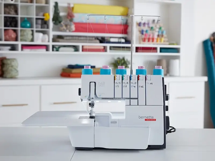 Overlock BERNETTE B68 AIRLOCK w kolorze białym z niebieskimi szpulkami, stojący na płaskim blacie. Widoczna funkcja renderki oraz stopki. Tło w jasnych kolorach, z różnorodnymi materiałami w plecaku.