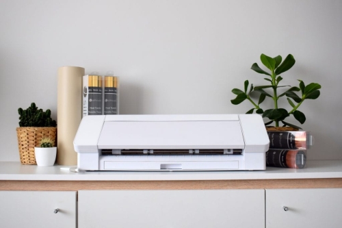 Ploter tnący Silhouette Cameo 4 w kolorze białym, umieszczony na białym blacie. W tle widoczne rolki materiałów do cięcia oraz rośliny doniczkowe. Urządzenie ma prostokątny kształt z widocznymi przyciskami i szczeliną tnącą.