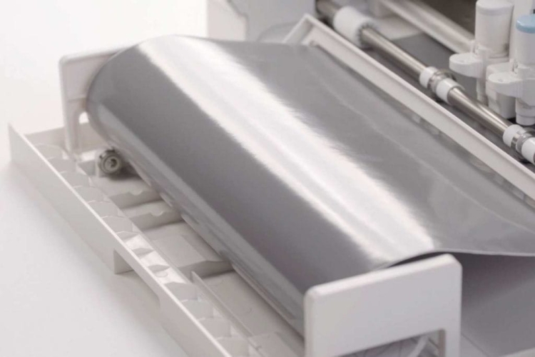 Ploter tnący Silhouette Cameo 4 w kolorze białym, z widoczną rolką błyszczącego, srebrnego materiału. Ujęcie z boku, pokazujące otwarte podajnik i elementy robocze urządzenia. Proste tło, które podkreśla detale maszyny.