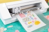 Ploter tnący Silhouette Cameo 4 w kolorze białym, z rolką papieru z wyciętymi kolorowymi naklejkami. Urządzenie widoczne z przodu, na zielonym tle, z detalami konstrukcyjnymi i elementami sterującymi na dolnym panelu.