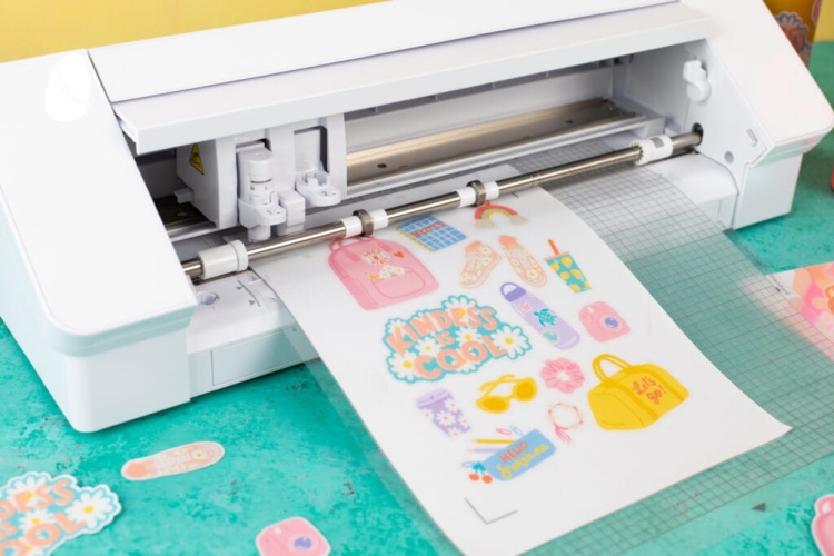Ploter tnący Silhouette Cameo 4 w kolorze białym, z rolką papieru z wyciętymi kolorowymi naklejkami. Urządzenie widoczne z przodu, na zielonym tle, z detalami konstrukcyjnymi i elementami sterującymi na dolnym panelu.