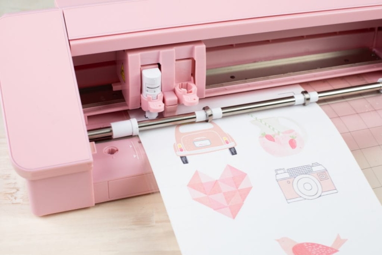 Różowy ploter tnący Silhouette Cameo 4 z widocznym ostrzem i rolką do cięcia. Na arkuszu papieru znajdują się wycięte grafiki: samochód, serce, aparat i owoc. Ujęcie z przodu, zbliżenie na detale urządzenia i cięcia.