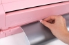 Ploter tnący Silhouette Cameo 4 w kolorze różowym, z widoczną ręką obsługującą mechanizm. Na zdjęciu widać srebrną folię wciąganą do urządzenia, które jest uchylone, ukazując jego górną część i detale konstrukcji.