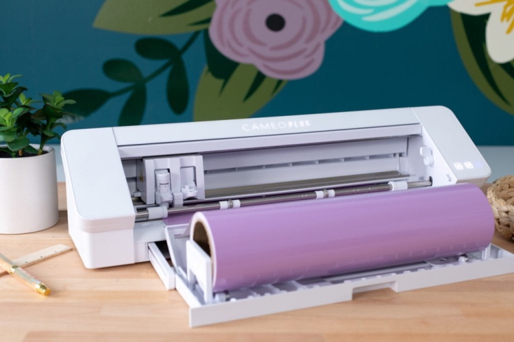 Ploter tnący Silhouette Cameo 4 Plus w kolorze białym, z rolką fioletowego materiału. Ujęcie z boku, pokazujące otwartą szufladę. W tle zielona ściana z kwiatowym motywem i doniczka z rośliną.