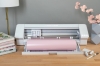 Ploter tnący Silhouette Cameo 4 Plus w kolorze białym z różowym rolką papieru, stojący na drewnianym blacie. W tle widoczne książki, narzędzia do craftingu oraz kwiat w doniczce. Otwarta komora z widocznymi głowicami tnącymi i przyciskami.