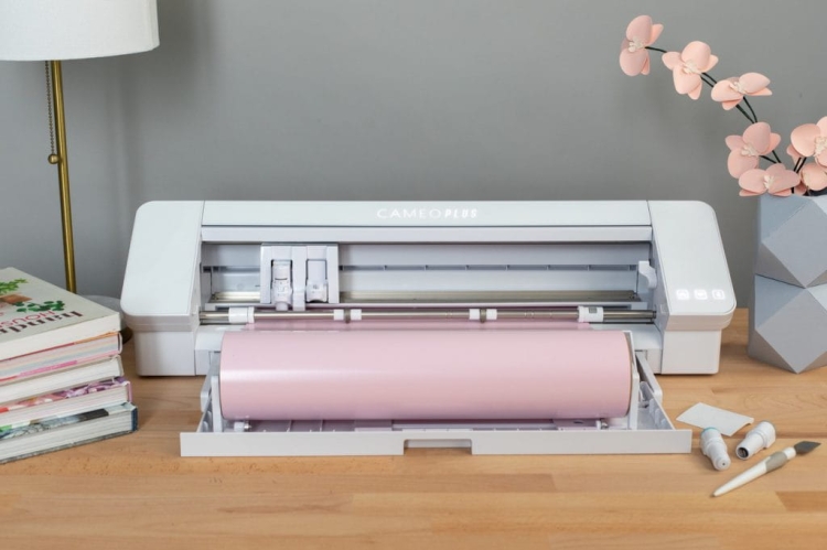 Ploter tnący Silhouette Cameo 4 Plus w kolorze białym z różowym rolką papieru, stojący na drewnianym blacie. W tle widoczne książki, narzędzia do craftingu oraz kwiat w doniczce. Otwarta komora z widocznymi głowicami tnącymi i przyciskami.