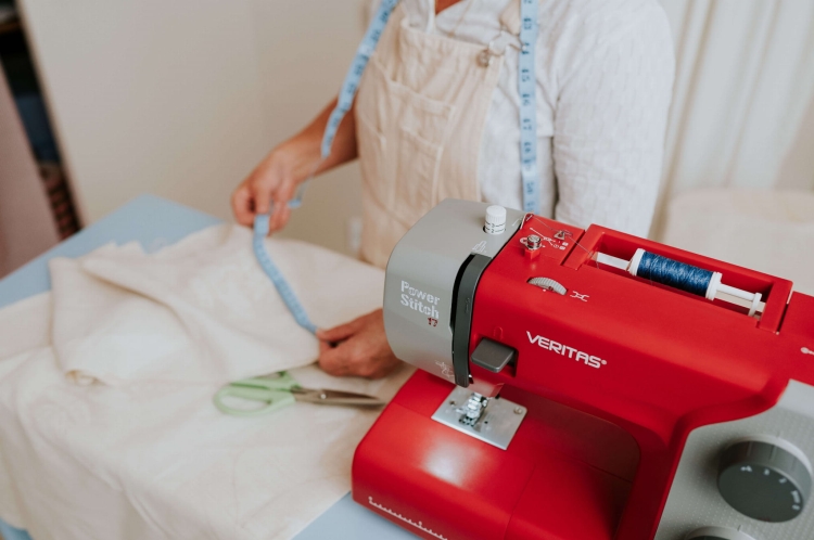 Maszyna do szycia Veritas Power Stitch 17 w kolorze czerwonym z 17 ściegami, widoczna z boku. Na stole leżą białe materiały oraz zielone nożyczki, a osoba w tle trzyma miarkę krawiecką.