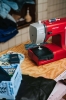 Maszyna do szycia Veritas Power Stitch 17 w kolorze czerwonym, ustawiona na drewnianym blacie. Widać czarny materiał pod stopką, a obok znajduje się niebieska miarka krawiecka oraz zestaw akcesoriów do szycia. W tle leżą kawałki materiału.