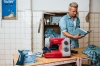 Maszyna do szycia Veritas Power Stitch 17 w kolorze czerwonym z szarymi detalami, z widocznymi pokrętłami do ustawienia ściegów. Ustawiona na stole, obok akcesoria do szycia i kawałek materiału w kratkę. W tle mężczyzna trzymający książkę.