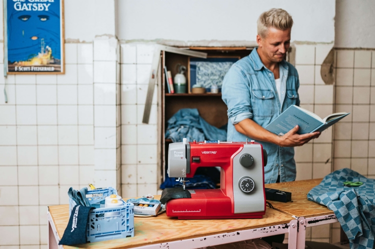 Maszyna do szycia Veritas Power Stitch 17 w kolorze czerwonym z szarymi detalami, z widocznymi pokrętłami do ustawienia ściegów. Ustawiona na stole, obok akcesoria do szycia i kawałek materiału w kratkę. W tle mężczyzna trzymający książkę.