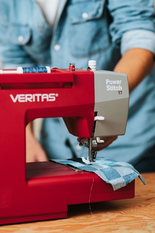 Maszyna do szycia Veritas Power Stitch 17 w kolorze czerwonym, z szarym panelem, prezentująca mechanizm szycia na niebieskiej tkaninie w kratę. Ujęcie z boku, z widocznym wątkiem i igłą, z ręką operatora trzymającą materiał.