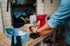 Maszyna do szycia Veritas Power Stitch 17 w kolorze czerwonym, umieszczona na drewnianym stole. Widoczne są czarne kawałki materiału oraz niebieski pojemnik z niciami. Osoba trzymająca materiał przygotowuje go do szycia, w tle widać niebieskie dżinsy.