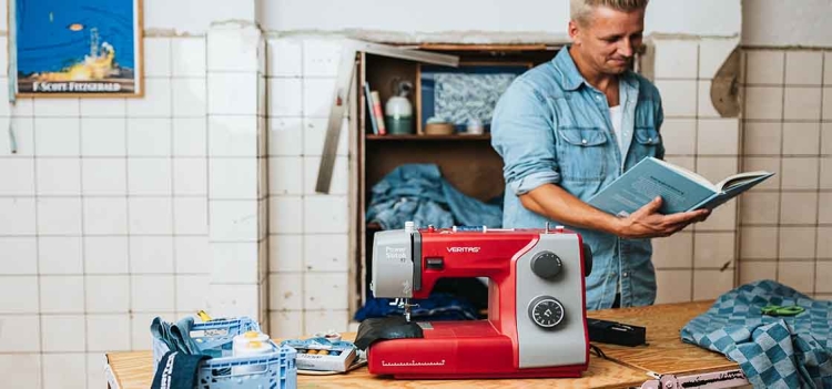 Maszyna do szycia Veritas Power Stitch 17 w kolorze czerwonym i szarym, stojąca na drewnianym stole. Widoczne pokrętło do wyboru ściegów oraz panel z napisem Power Stitch. W tle niebieskie materiały oraz mężczyzna trzymający książkę.