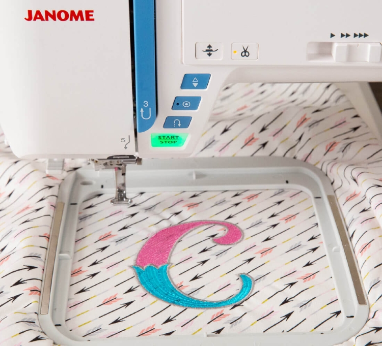 Maszyno-hafciarka Janome Skyline S9 w zbliżeniu, z widocznym haftem litery C w różowo-niebieskich kolorach. Ujęcie pokazuje panel sterujący z przyciskiem START/STOP oraz igłę na tle materiału w strzałki.